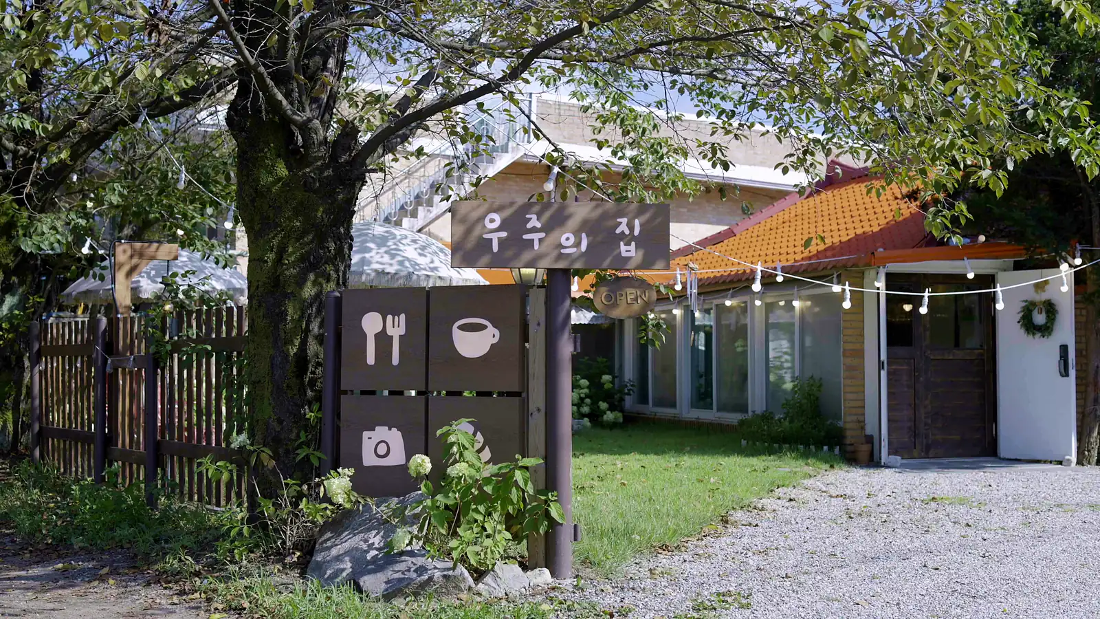 경기 용인 처인구 양지읍의 카페 우주의 집 외관. 자갈길과 나무 울타리, 기와지붕과 전구 스트링이 이어진다.
