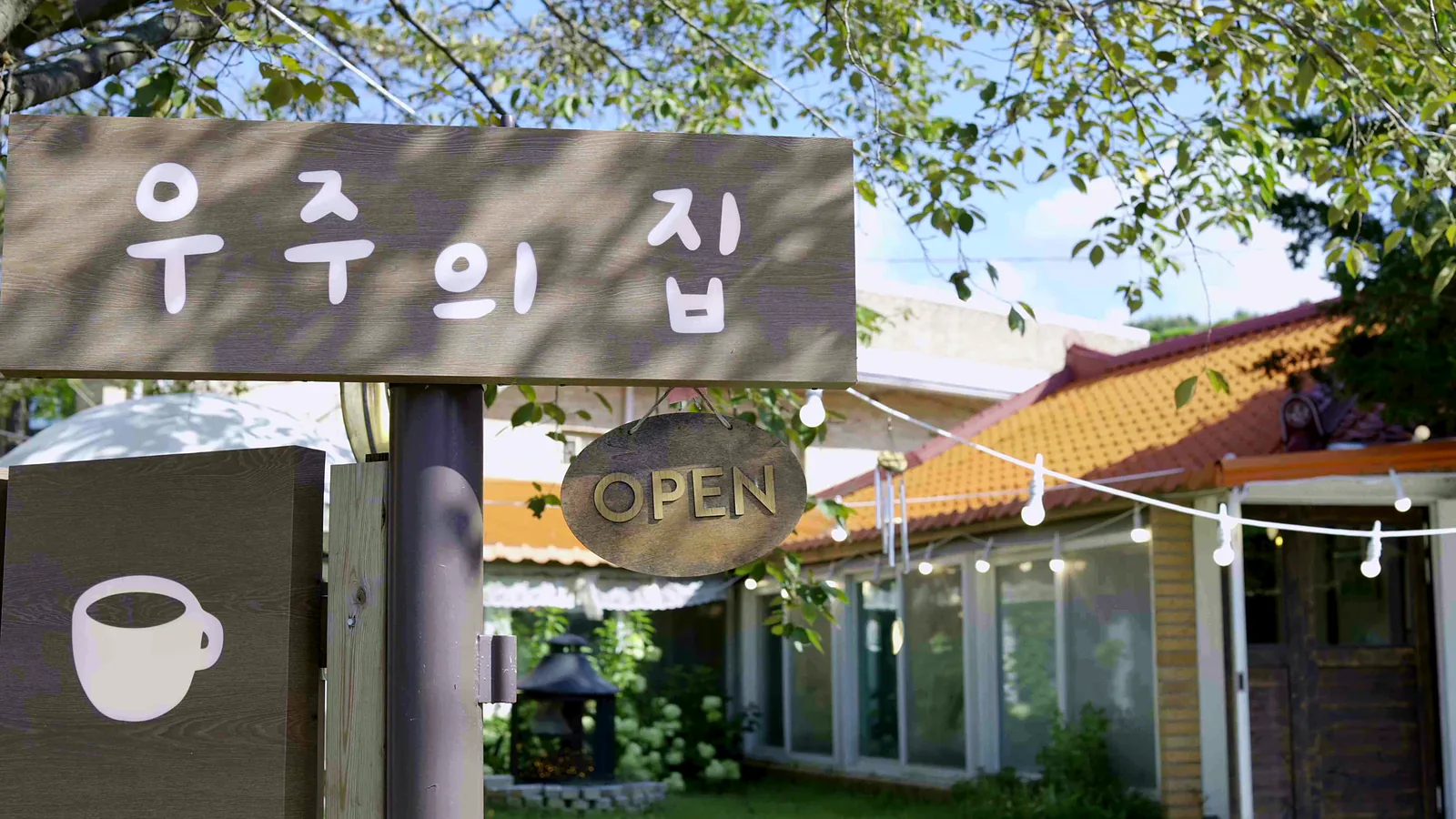 ‘우주의 집’이라고 적힌 나무 간판과 OPEN 사인, 커피잔 그림 입간판. 뒤로 주황 기와지붕이 보인다.